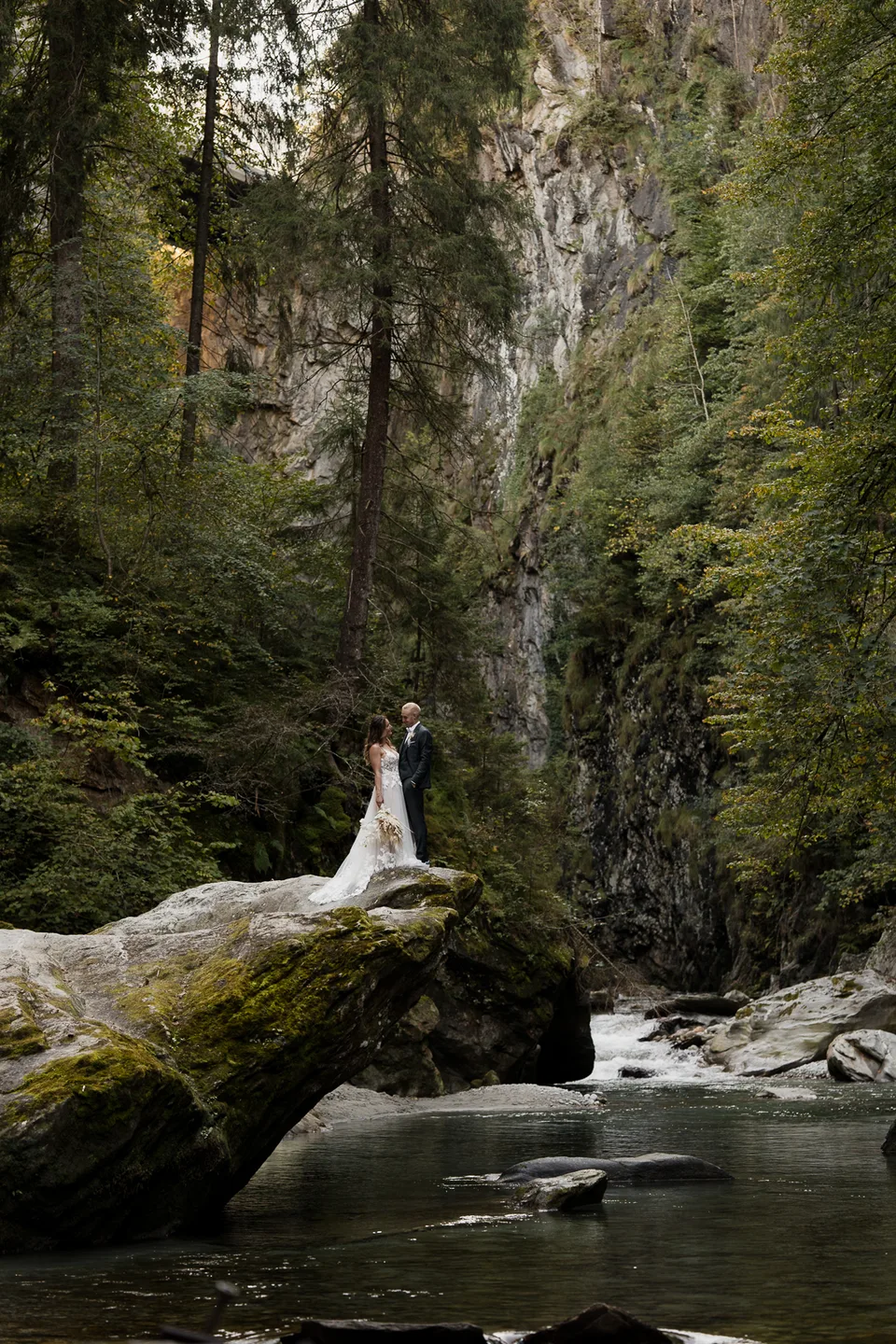 Braut steht im Brautkleid auf einem Felsen in einer bewaldeten Schlucht am Wasser