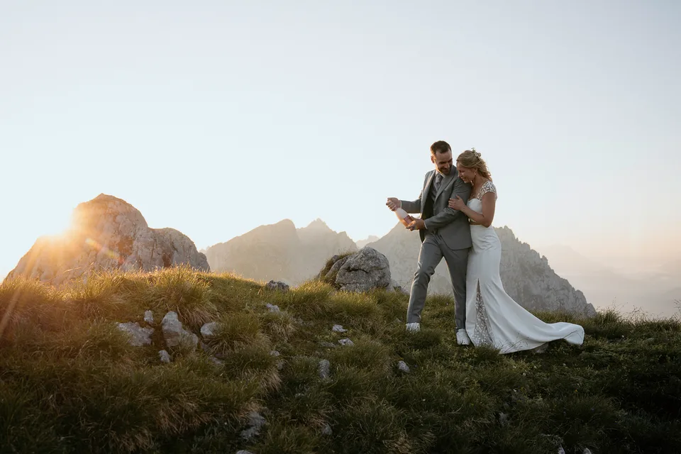 Brautpaar umarmt sich auf einer Bergwiese, im Hintergrund Gipfel im Licht der aufgehenden Sonne
